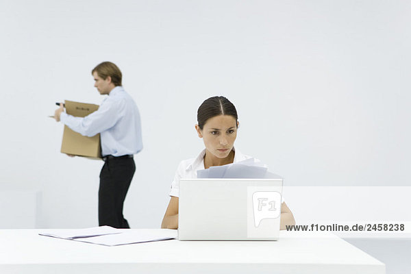 Professionelle Frau beim Durchsehen von Dokumenten am Schreibtisch hinter dem Laptop  Kollegin mit Box im Hintergrund