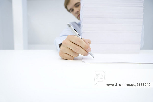 Mann sitzt hinter einem hohen Stapel von Dokumenten und schreibt auf ein Blatt Papier.