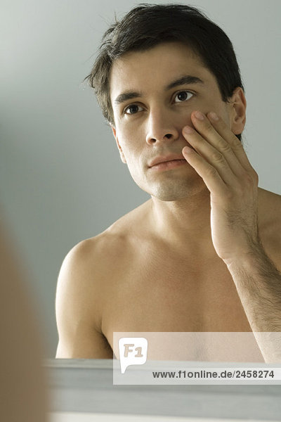 Nackter Mann  der sich selbst im Spiegel ansieht und sein Gesicht berührt.