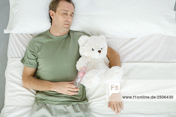 Father asleep in bed  holding baby bottle and teddy bear  overhead view