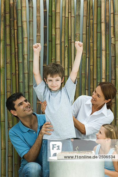 Kleiner Junge neben Topfkeimling stehend  Arme erhoben  Familie lächelnd