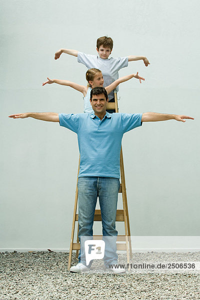 Familie steht auf einer Leiter mit ausgestreckten Armen  Mädchen schaut weg  Vater und Sohn lächeln in die Kamera.