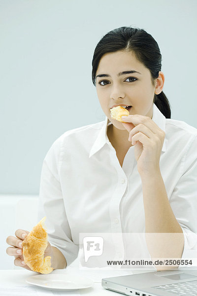 Junge Frau isst Croissant  sitzt auf dem Schreibtisch  schaut in die Kamera.