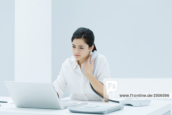 Junge berufstätige Frau am Schreibtisch sitzend  mit Laptop  Festnetztelefon an der Brust haltend