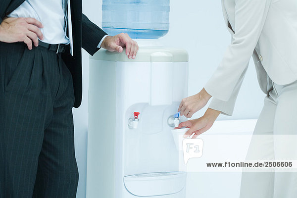 Professionelle Frau füllt Einwegbecher mit Wasser aus dem Wasserkühler  männlicher Kollege steht in der Nähe  Ausschnittansicht