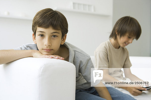 Jungen sitzen auf der Couch  einer schaut in die Kamera  der andere spielt Handheld-Videospiel