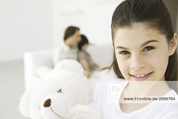 Mädchen mit Teddybär  Familie im Hintergrund auf der Couch sitzend