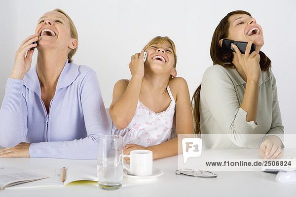 Zwei Frauen und ein junges Mädchen am Tisch  jede mit dem Handy  alle lachend und nach oben schauend.