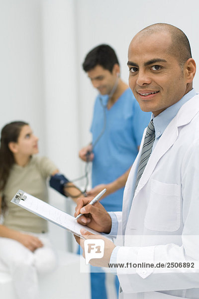 Arzt schreibt auf Klemmbrett  lächelt in die Kamera  Krankenschwester misst den Blutdruck des Patienten im Hintergrund
