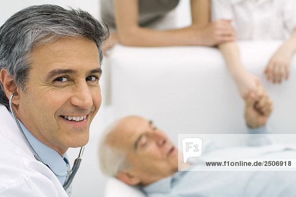 Doctor smiling at camera  senior man lying on couch in background
