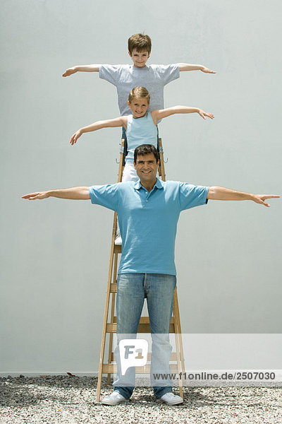 Junge und Mädchen stehen auf einer Leiter mit den Armen in der Luft  Vater steht auf dem Boden  alle lächeln in die Kamera.