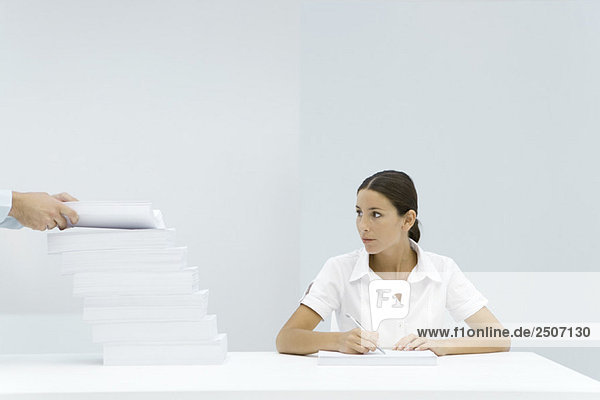 Frau sitzt am Tisch  schaut auf einen gestaffelten Stapel Papier  die Hände des Mannes fügen dem Stapel Papier hinzu.