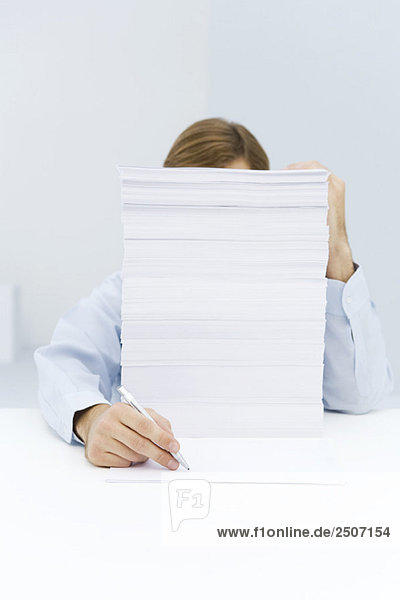 Ein Mann  der sich hinter einem hohen Stapel Papier versteckt  um auf ein einziges Blatt Papier zu schreiben.
