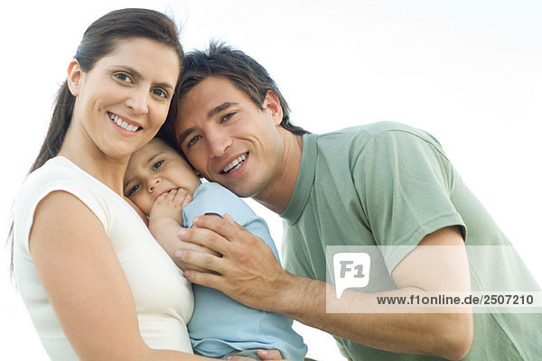 Familie lächelt in die Kamera  Mutter hält Baby-Tochter  Vater lehnt sich hinein und umarmt das Baby sanft.
