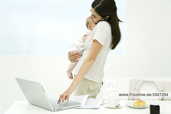 Professional woman standing beside breakfast table  holding baby  using cell phone and laptop computer