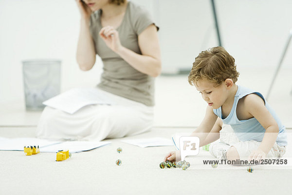 Kleiner Junge auf dem Boden sitzend  Murmeln spielend  Mutter von Papierkram umgeben im Hintergrund