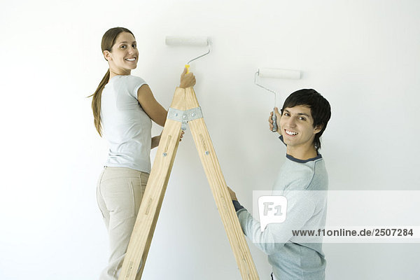 Couple painting wall with rollers  both smiling over their shoulder at camera