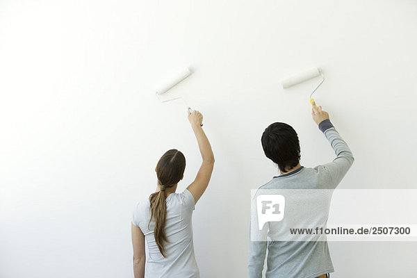 Couple using paint rollers on wall  rear view