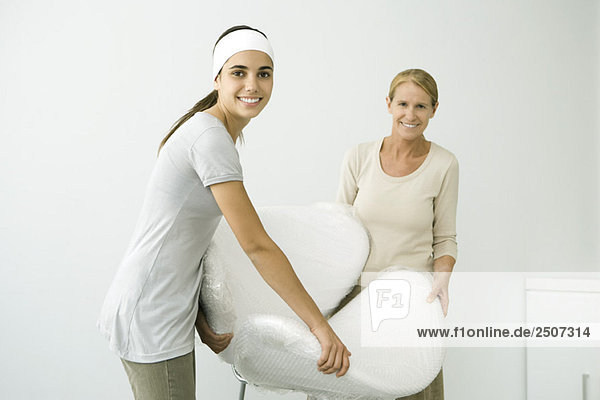 Mother and daughter carrying chair covered in bubble wrap  smiling at camera