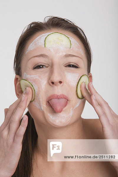 Junge Frau  die eine Gurken-Gesichtsmaske aufträgt und ihre Zunge herausstreckt.