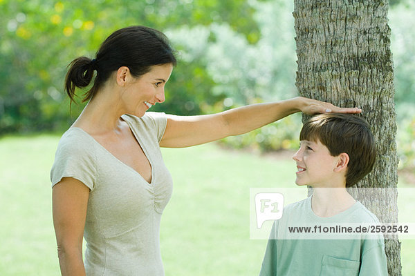 Boy standing against tree trunk  his mother placing her hand on his head  both smiling looking at each other