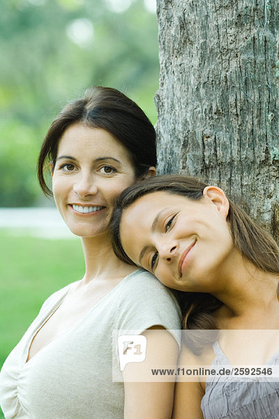 Mutter und Teenagertochter lehnen sich an den Baum  Mädchenkopf auf der Schulter der Mutter  lächelnd