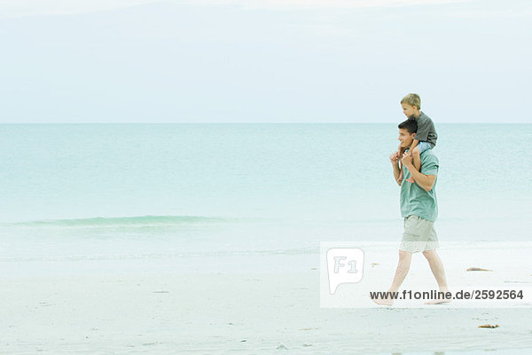 Man walking on empty beach carrying son on his shoulders