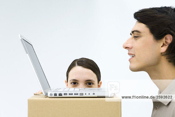 Mann mit Laptop am Kartonschreibtisch  Frau versteckt sich hinter der Kiste  schaut über die Kante