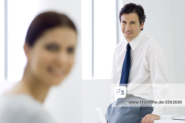Professional man leaning against desk  smiling at camera  female colleague in foreground