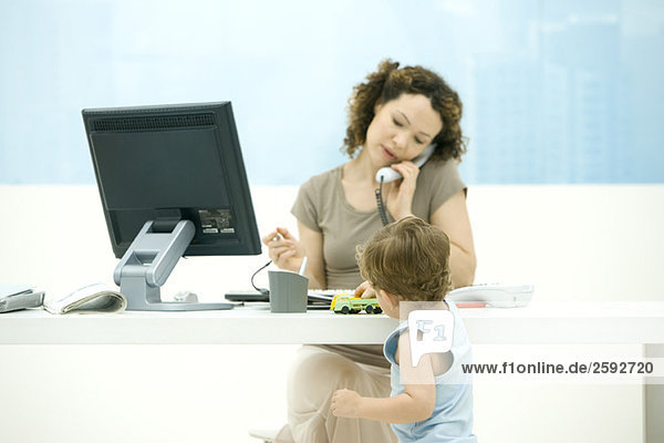 Frau mit Telefon im Büro  Kleinkind Sohn spielt mit Spielzeug LKW auf dem Schreibtisch