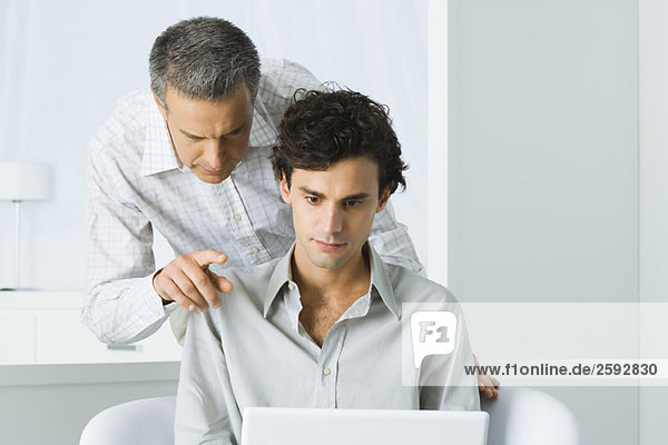 Mature man leaning over young man's shoulder  both looking at laptop computer