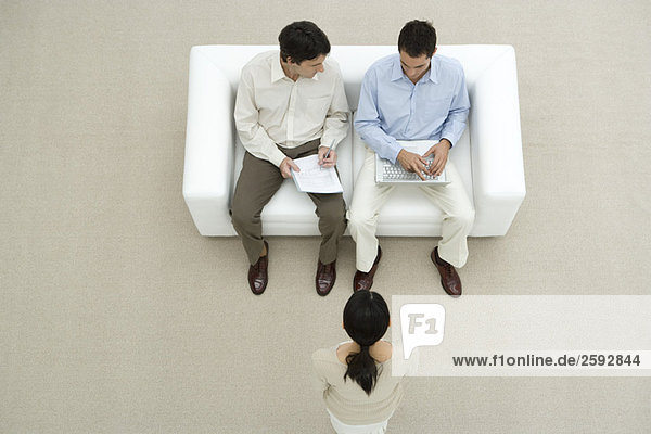 Professionelle Männer auf dem Sofa sitzend  diskutierendes Dokument  Frau vor ihnen stehend  Blick über Kopf