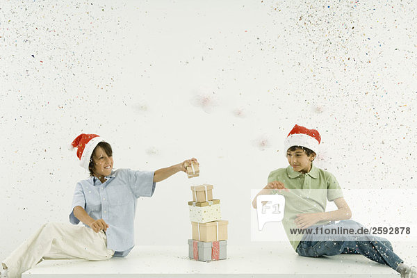 Zwei Jungen mit Weihnachtsmützen stapeln Geschenke  Konfetti fallen um sie herum.