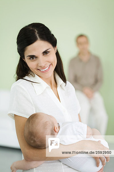 Mother holding baby  smiling at camera  man in background