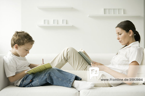 Mutter und Sohn sitzen von Angesicht zu Angesicht auf dem Sofa  Frau liest  Junge schreibt im Buch