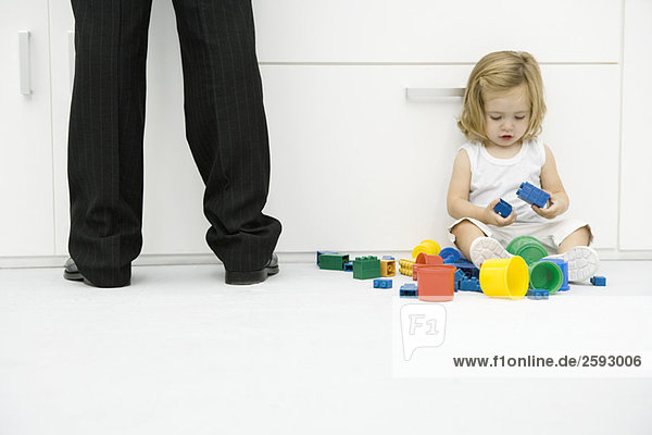Kleinkind auf dem Boden neben Vaters Beinen sitzend  mit Spielzeug spielend