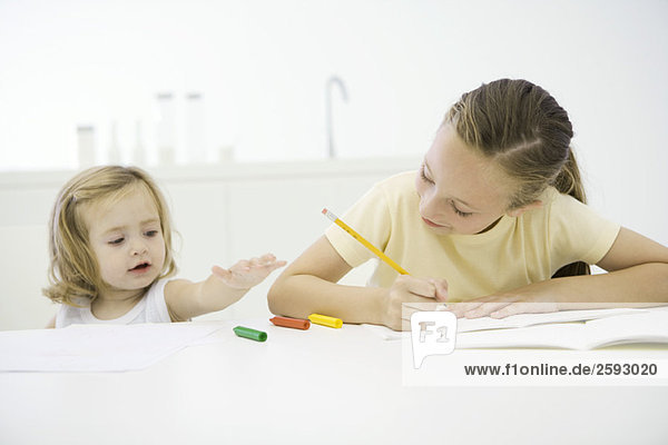 Mädchen macht Hausaufgaben am Tisch  jüngere Schwester greift nach Buntstiften