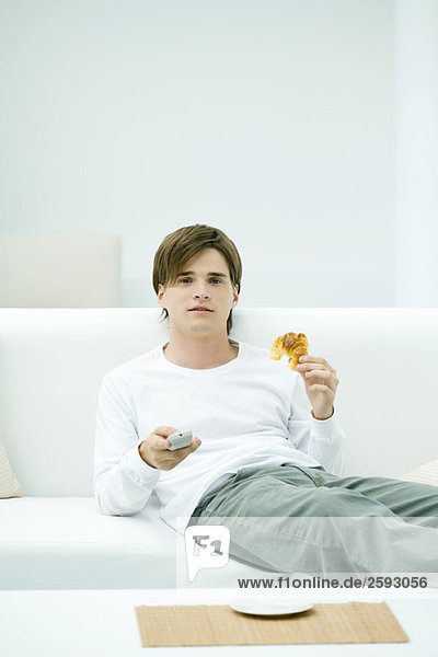 Junger Mann sitzt auf dem Sofa und hält Fernbedienung und Croissant.