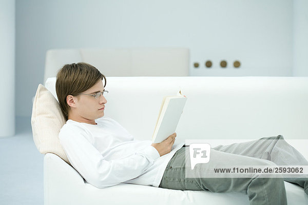 Junger Mann auf Sofa liegend  Lesebuch  Seitenansicht