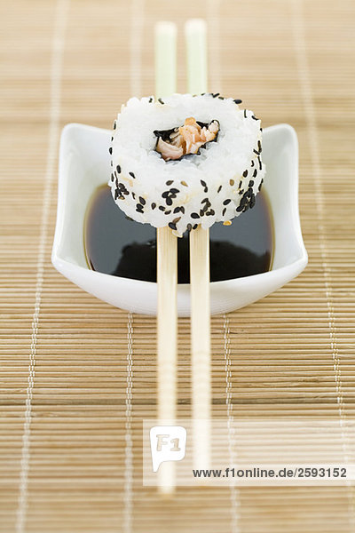 Maki sushi balanced on chopsticks over dish of soy sauce