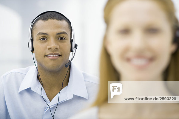 Professional man wearing headset smiling at camera  female colleague in foreground