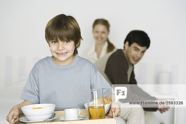 Kleiner Junge mit Frühstück auf Tablett  Eltern sitzen im Hintergrund auf dem Bett.