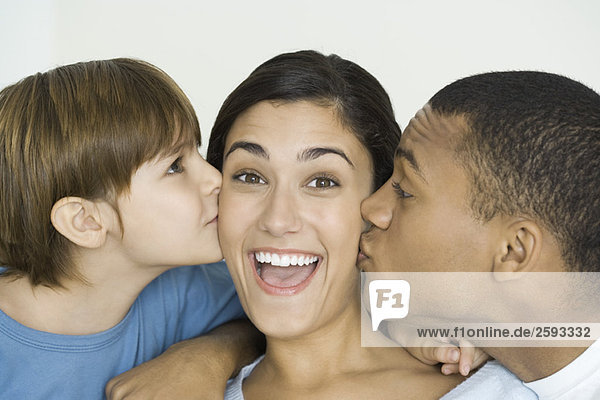 Frau lächelt vor der Kamera als Mann und Sohn ihre Wangen küssen