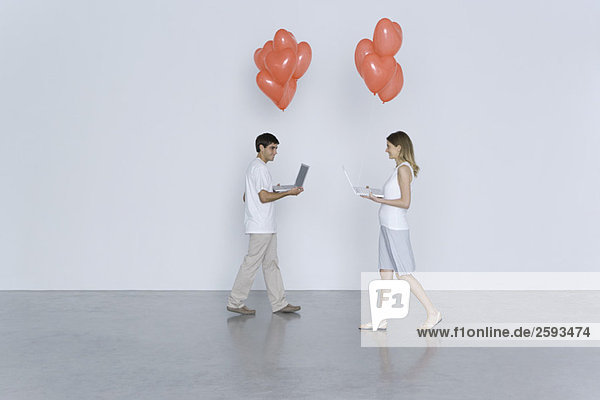 Young man and woman walking toward each other  both carrying laptop computers and heart balloons