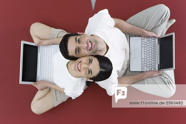 Paar sitzt Rücken an Rücken auf dem Boden  hält Laptop-Computer und lächelt vor der Kamera.