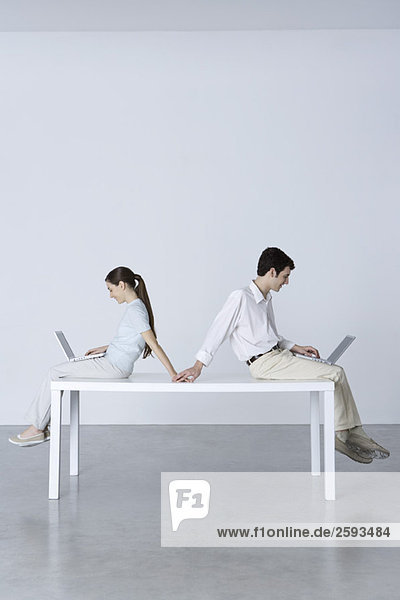 Paar sitzt Rücken an Rücken  benutzt Laptops und hält sich an den Händen.