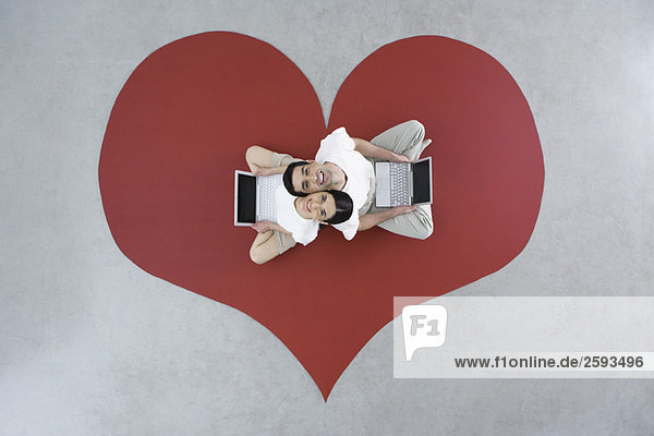 Couple sitting back to back on large heart  holding laptop computers  smiling up at camera