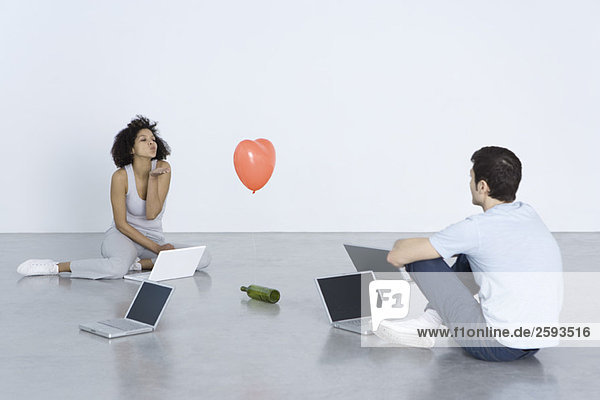Mann und Frau sitzen mit Laptops  Frau bläst Mann einen Kuss  Flasche und Herzballon dazwischen