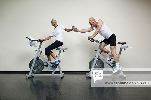 A senior woman and a mature man riding stationary bikes and passing a water bottle