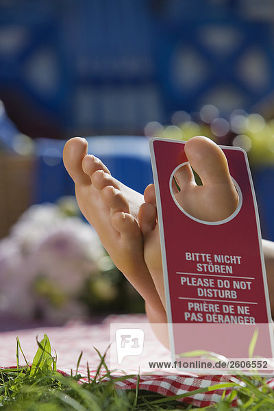 'Frau liegt auf Decke in der Wiese mit Schild ''Bitte nicht stören'' am Zehen Picknickkorb im Hintergrund Nahaufnahme von Füßen'